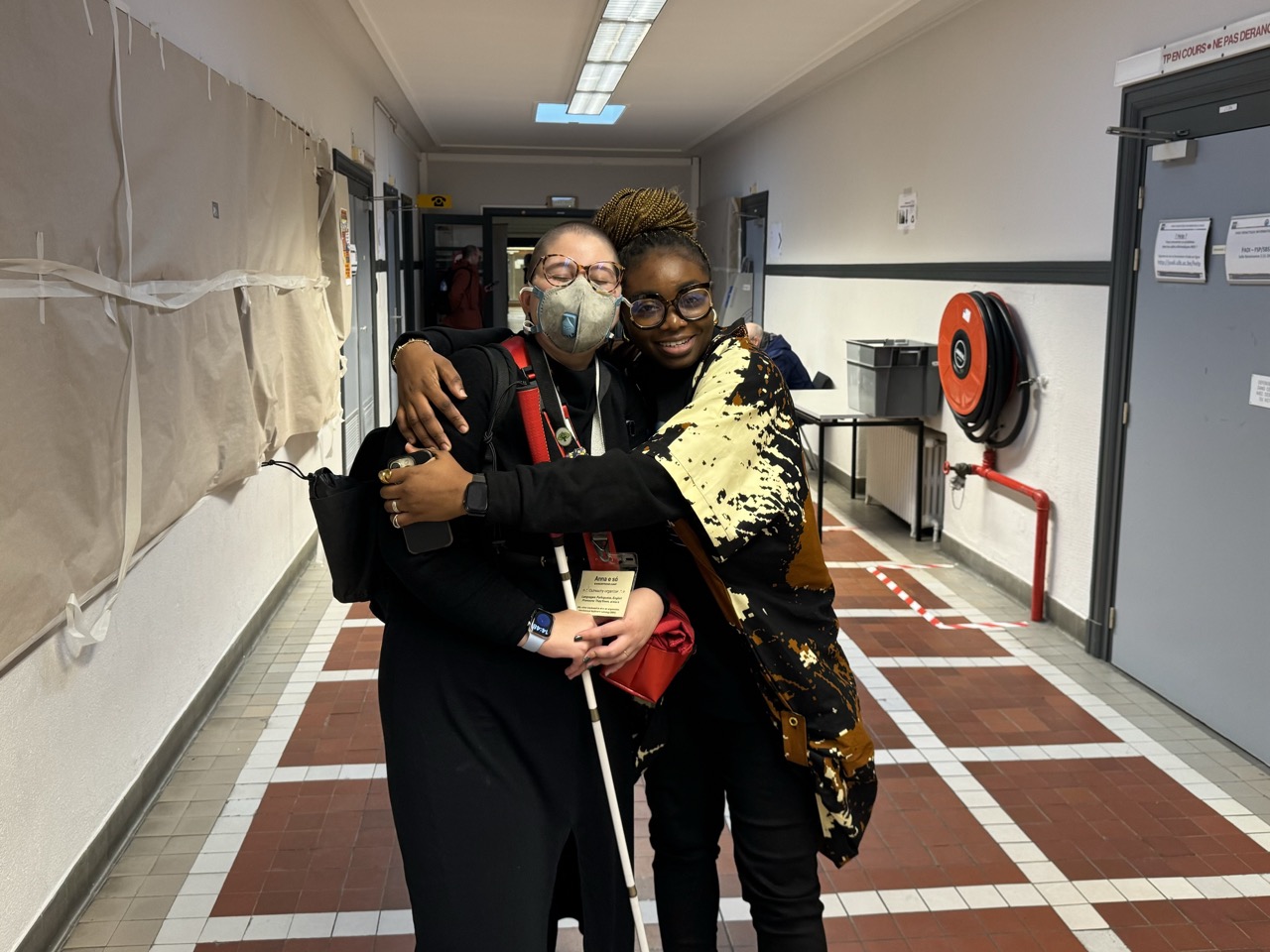 A photograph of Anna e só, a white femme person with a shaved head, being hugged in a hallway at ULB by Omotola Omotayo, a Black woman with an elegant golden brown braided hair in a bun. We're both wearing prescription glasses and predominantly black clothing. I'm wearing an N95 mask and carrying a distinctive bright red purse and a long white cane; Omotola is wearing an African gown with patterns in cream white and brown.